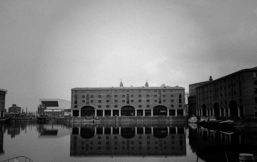 A black and white photo of the Liverpool Docks on a clear day showing a reflection of the brick dockland buildings in the water